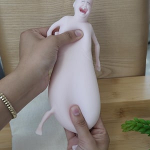 May include: A light pink, abstract sculpture of a crying baby with a bulbous body and small limbs. The sculpture is held by two hands, and has a small tuft of hair on its head.