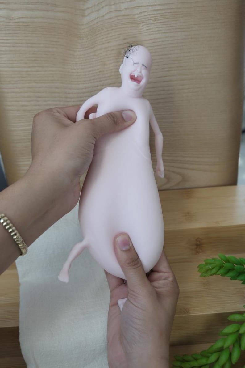 May include: A light pink, abstract sculpture of a crying baby with a bulbous body and small limbs. The sculpture is held by two hands, and has a small tuft of hair on its head.