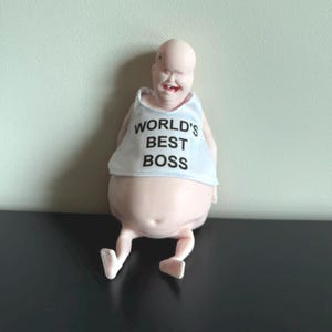 May include: A novelty figurine of a bald, smiling figure with a white tank top. The tank top reads "WORLD'S BEST BOSS" in black text. The figure is light pink and has small legs and feet.