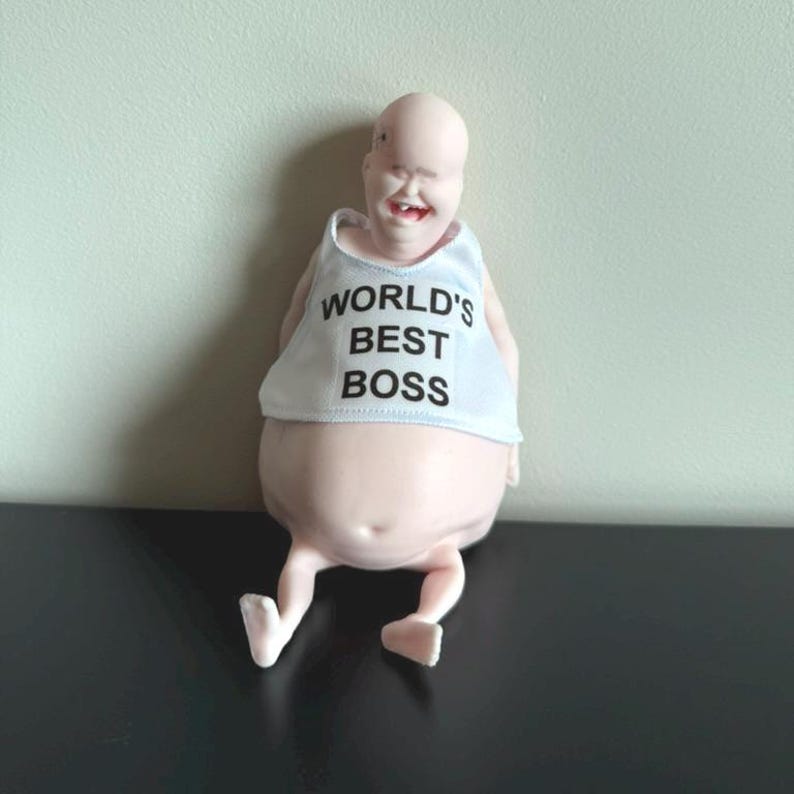 May include: A novelty figurine of a bald, smiling figure with a white tank top. The tank top reads "WORLD'S BEST BOSS" in black text. The figure is light pink and has small legs and feet.
