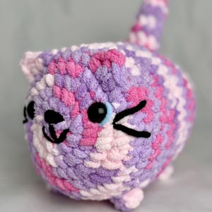 May include: A handmade, round crochet cat in shades of purple, pink, and white. The cat has black eyes, a winking eye, and a smiling mouth. The cat has a small tail and small feet.