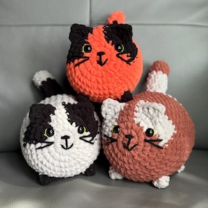 May include: Three handmade crochet cat plushies in orange, white, black, and brown. Each plush has a round body, small ears, and a smiling face with black embroidered details. The cats are arranged in a playful stack.