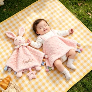Puede incluir: Un bebé yace sobre una manta a cuadros amarillos junto a una manta de seguridad rosa y un conejito de peluche. La manta de seguridad es rosa con una textura punteada y el nombre "Amelia" bordado. El conejito tiene orejas rosas y un cuello estampado colorido.