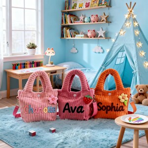 Puede incluir: Tres bolsos tote de pana en rosa y naranja, cada uno bordado con un nombre: "Charlotte", "Ava" y "Sophia". Los bolsos tienen correas a juego y detalles decorativos. Están sobre una alfombra azul en una habitación infantil.