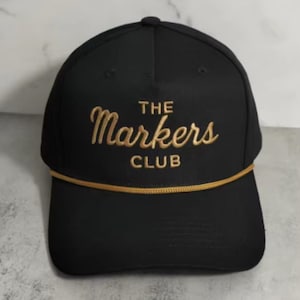Może przedstawiać: Czarna czapka bejsbolowa z napisem "THE Markers CLUB" haftowanym złotą nicią. Czapka ma strukturalną koronę, zakrzywiony daszek i złoty sznurek.