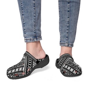 May include: Black and white patterned clogs. The clogs feature a geometric design with a strap and a molded footbed. The shoes are worn with blue jeans. The clogs are a slip-on style.