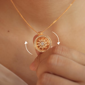 Peut inclure: Un pendentif circulaire doré avec un motif floral central incrusté de petites pierres claires. Le pendentif est entouré d'une bordure de minuscules pierres et est suspendu à une fine chaîne dorée. Des flèches blanches courbes indiquent la rotation.