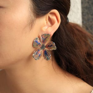 May include: A large, five-petaled flower earring featuring a textured, metallic silver finish with streaks of blue, red, and gold paint. The earring has a prominent, organic shape with irregular edges and a dark center, worn on the earlobe.