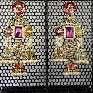 May include: A pair of ornate gold-tone dangle earrings featuring a central rectangular purple stone, circular red and green enamel accents, clear rhinestones, and multiple small bell-shaped charms adorned with tiny white beads and red enamel.