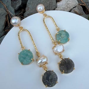 Può includere: Orecchini pendenti color oro con borchie di perle, dettagli dorati curvi e pietre preziose. Gli orecchini presentano una pietra verde-azzurra chiara, una perla bianca e una pietra grigio scuro. Gli orecchini sono esposti su una superficie bianca.