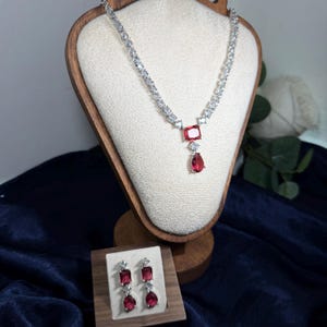 Può includere: Un set di collana e orecchini argentati. La collana presenta una serie di piccole pietre trasparenti e un pendente con una grande pietra rossa quadrata e una pietra rossa a forma di goccia. Gli orecchini hanno un design simile, con una pietra rossa quadrata e a goccia.