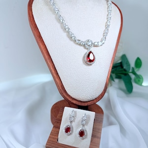 Könnte beinhalten: Ein silberfarbenes Halsketten- und Ohrring-Set. Die Halskette hat eine Reihe klarer, funkelnder Steine und einen tropfenförmigen roten Edelsteinanhänger. Die Ohrringe haben ebenfalls rote, tropfenförmige Edelsteine. Der Schmuck wird auf einem Holzständer präsentiert.