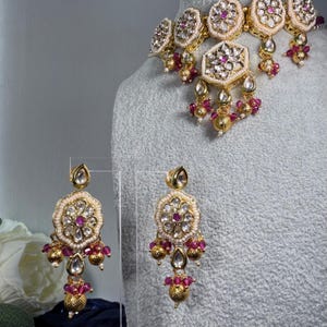 Könnte beinhalten: Ein goldfarbenes Halsketten- und Ohrring-Set. Der Schmuck weist komplizierte Blumen- und geometrische Designs mit klaren und rubinfarbenen Edelsteinen sowie kleine goldene Perlen auf. Die Halskette wird auf einer Schaufensterpuppe präsentiert.