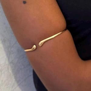 May include: A gold-colored arm cuff with a minimalist design. The open cuff features teardrop-shaped ends, creating a sleek and modern aesthetic. The jewelry is worn on the arm, showcasing its elegant simplicity.