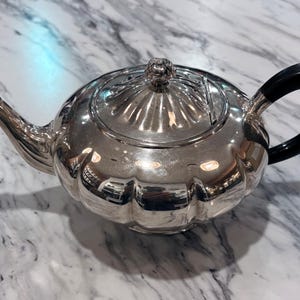May include: A silver teapot with a black handle and a decorative lid. The teapot has a fluted body and a curved spout. The handle is a rounded, semi-circular shape. The lid has a small, decorative knob.