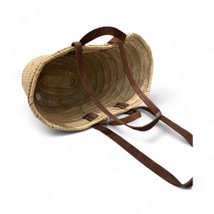 May include: A woven straw tote bag with brown leather straps. The bag has a rounded shape and a natural color, with the leather straps attached at the top and sides. The straps are long and appear to be adjustable.