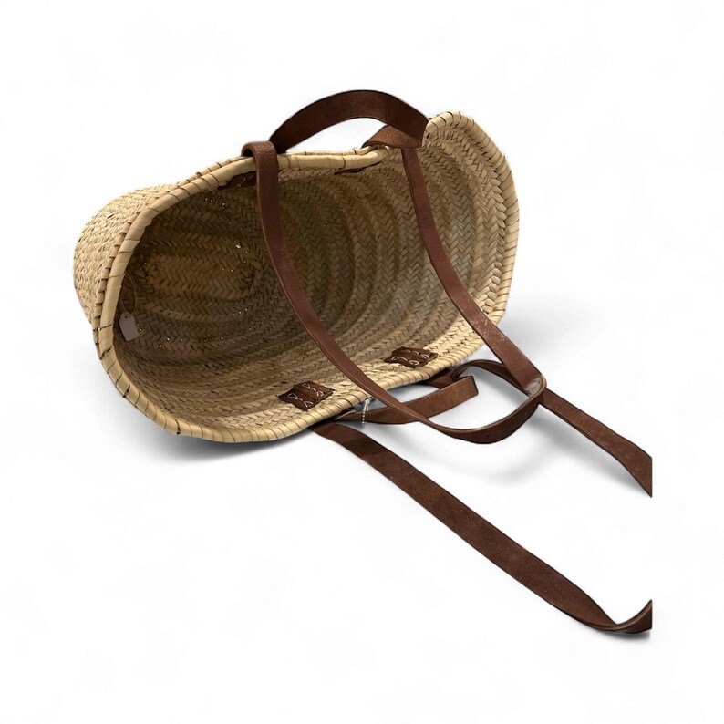 May include: A woven straw tote bag with brown leather straps. The bag has a rounded shape and a natural color, with the leather straps attached at the top and sides. The straps are long and appear to be adjustable.