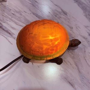 May include: A turtle-shaped lamp with an orange, textured shell and dark brown legs and head. The shell glows with a warm light, and the lamp has a black power cord. The lamp is on a white and gray marble surface.