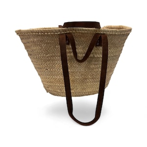 May include: Woven straw tote bag with brown leather handles. The bag has a wide, open top and a sturdy, rectangular base. The handles are attached to the bag with metal rivets. The bag is designed for carrying items.