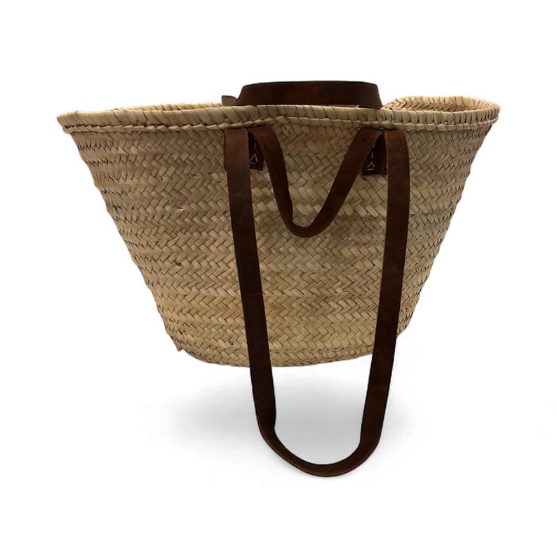 May include: Woven straw tote bag with brown leather handles. The bag has a wide, open top and a sturdy, rectangular base. The handles are attached to the bag with metal rivets. The bag is designed for carrying items.