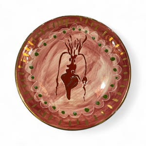 May include: A round, hand-painted ceramic plate with a maroon and white background. The plate features a dark brown abstract design in the center, a scalloped border with green dots, and a gold rim. The plate is a decorative piece.
