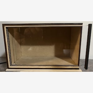Puede incluir: Una vitrina rectangular de madera con frente de cristal transparente y puerta corredera. El marco es de color marrón oscuro y el interior es de color beige claro. La vitrina se apoya sobre una base clara.