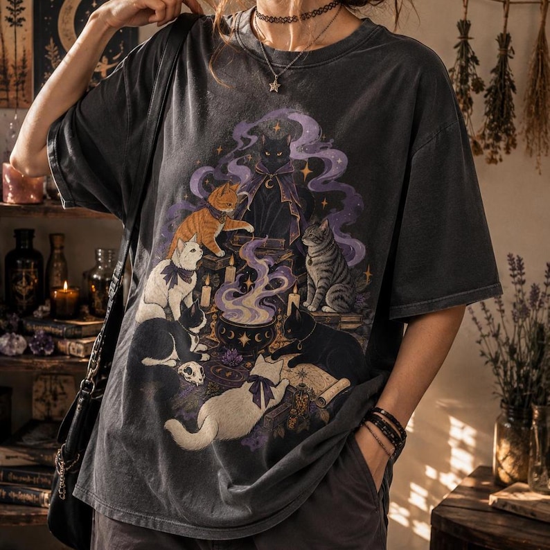 May include: A charcoal gray short-sleeve t-shirt featuring a graphic print of six cats surrounding a steaming cauldron. The illustration includes a black cat in a purple cloak, an orange tabby, a white cat, a gray tabby, and two black cats.
