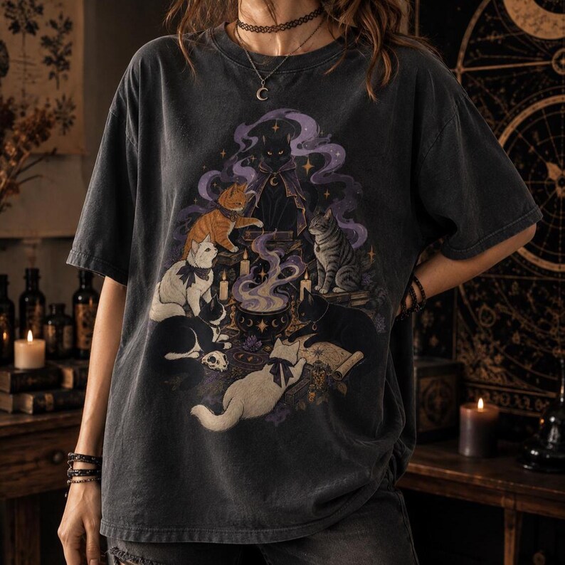May include: A charcoal gray short-sleeve t-shirt featuring a graphic print of several cats gathered around a steaming cauldron. The illustration includes a black cat in a cape, an orange tabby, a white cat, and a gray striped cat amidst purple smoke.