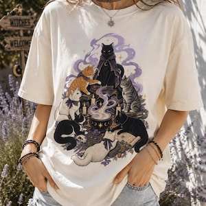 May include: An off-white short-sleeve t-shirt featuring a graphic illustration of seven cats gathered around a central cauldron emitting purple smoke. The cats are depicted in various colors including black, white, orange, and tabby, with some wearing small capes or bows.