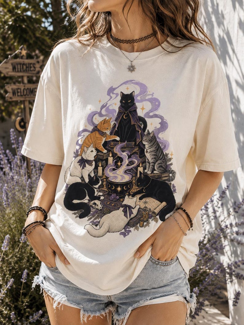 May include: An off-white short-sleeve t-shirt featuring a graphic illustration of seven cats gathered around a central cauldron emitting purple smoke. The cats are depicted in various colors including black, white, orange, and tabby, with some wearing small capes or bows.
