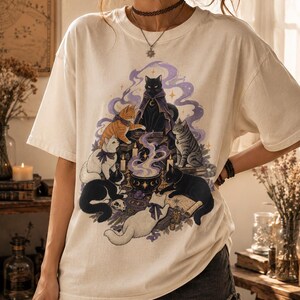 May include: An off-white short-sleeve t-shirt featuring a graphic print of six cats gathered around a steaming cauldron. The illustration includes a black cat in a purple cloak, an orange tabby, a white cat, a gray tabby, and two black cats.