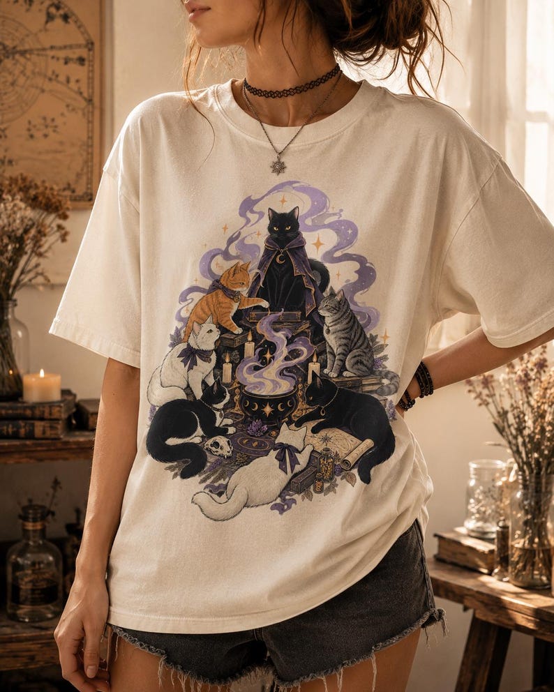 May include: An off-white short-sleeve t-shirt featuring a graphic print of six cats gathered around a steaming cauldron. The illustration includes a black cat in a purple cloak, an orange tabby, a white cat, a gray tabby, and two black cats.