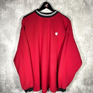 May include: A red sweatshirt with a white Apple logo on the chest. The collar and cuffs have black and white striped detailing. The sweatshirt is hanging on a black hanger against a gray background.