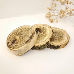 May include: Three wooden coasters made from tree slices. The coasters have a natural wood grain pattern with concentric circles and a light brown color. The edges have a rough bark texture.