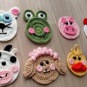 Può includere: Una collezione di sottobicchieri fatti a mano all'uncinetto con facce di animali, tra cui un gatto bianco, una rana verde, un maiale rosa, una gallina bianca, una mucca bianca e nera, una pecora marrone, un'anatra gialla e un cane marrone. Ogni sottobicchiere ha dettagli.