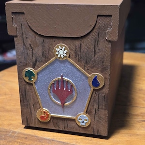 May include: A brown wooden box with a lid, featuring a decorative emblem with a red hand symbol at the center. The emblem is surrounded by gold trim and symbols representing different elements. The box is designed for storing items.