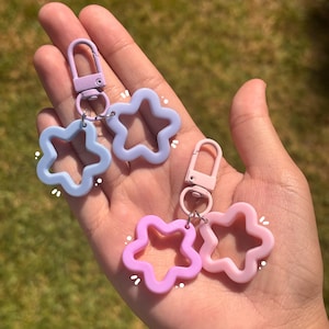 May include: Two keychains featuring open star-shaped charms. One keychain has a light purple clasp with two periwinkle blue star charms, and the other has a light pink clasp with two pink star charms. Each charm has small white graphic accents.