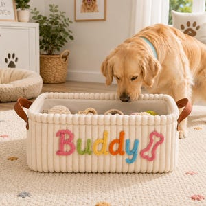 May include: A cream-colored, corduroy dog toy basket with the name "Buddy" in colorful letters. The basket has brown leather handles and is filled with various dog toys. A golden retriever is sniffing the basket.