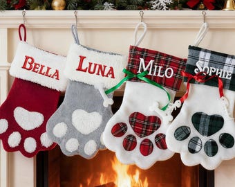 May include: Four Christmas stockings shaped like paw prints hang above a fireplace. Each stocking is personalized with a name: Bella, Luna, Milo, and Sophie. The stockings have red, gray, and white accents.