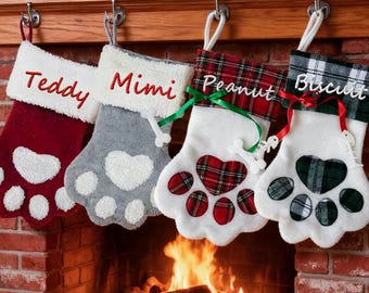May include: Four Christmas stockings shaped like pet paws hang above a brick fireplace. Each stocking is a different color: red, gray, white, and green. The stockings are personalized with the names "Teddy", "Mimi", "Peanut", and "Biscuit".