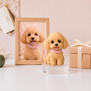 May include: A tan-colored dog figurine with a pink collar sits on a clear stand. A framed photo of a similar dog is behind it. A small gift box with a ribbon is to the right. The background is a light pink.