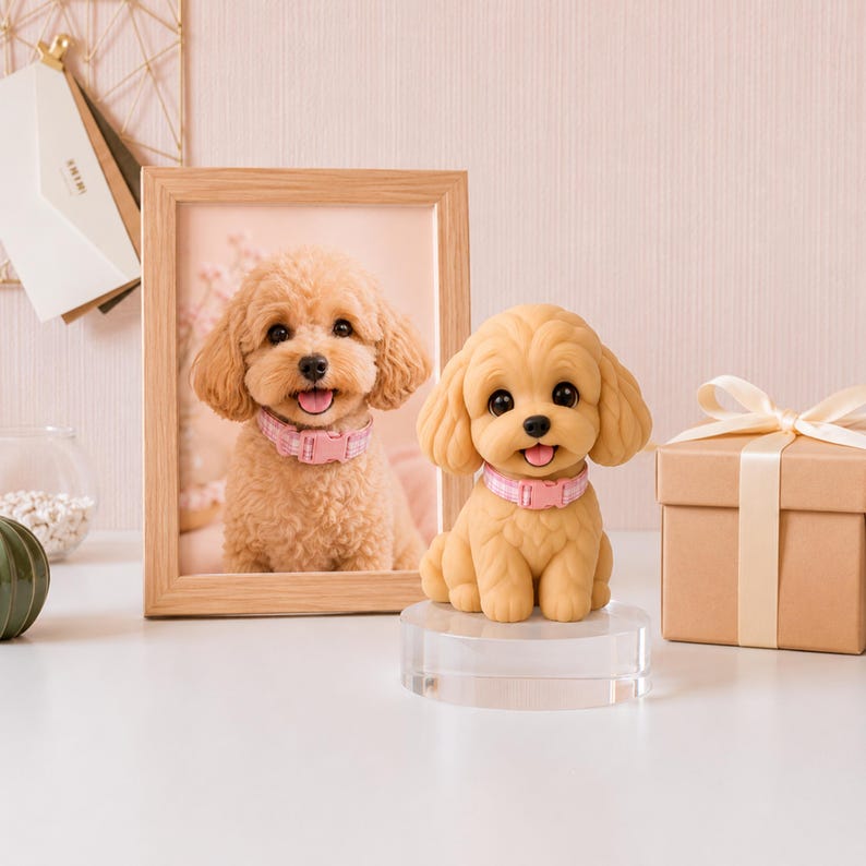 May include: A tan-colored dog figurine with a pink collar sits on a clear stand. A framed photo of a similar dog is behind it. A small gift box with a ribbon is to the right. The background is a light pink.
