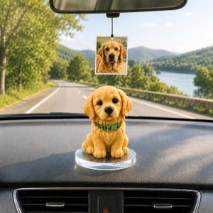 May include: A golden retriever puppy figurine with a green collar sits on a clear, round base on a car dashboard. A photo of a golden retriever hangs from the rearview mirror. The background shows a scenic road and a lake.