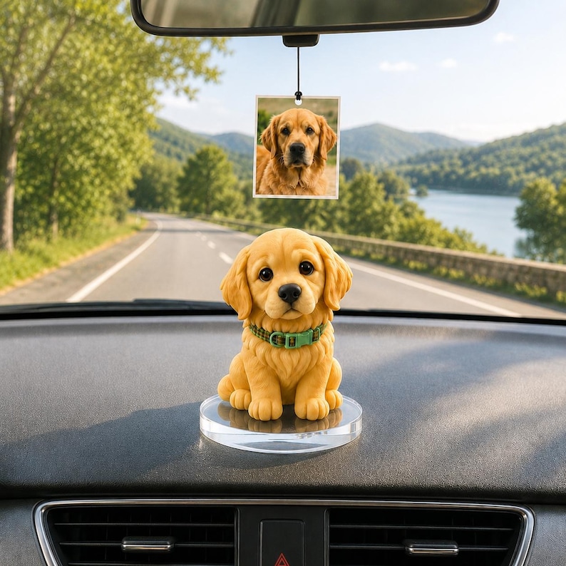 May include: A golden retriever puppy figurine with a green collar sits on a clear, round base on a car dashboard. A photo of a golden retriever hangs from the rearview mirror. The background shows a scenic road and a lake.