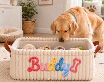 May include: A rectangular cream-colored storage basket with a ribbed texture and brown leather side handles. The front features the name Buddy in colorful knitted script letters, followed by a pink knitted bone shape, filled with various toys.