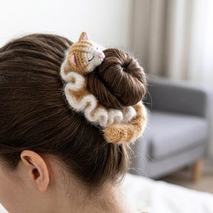 May include: A handmade crochet scrunchie with a sleeping cat design. The cat is tan and white, with pink accents on its ears. The scrunchie is wrapped around a bun of brown hair, creating a unique and whimsical hair accessory.