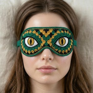 May include: A green, black, and gold crochet sleep mask with large, stylized yellow eyes. The mask is designed to cover the eyes and is made of yarn. The sleep mask is designed to block out light.
