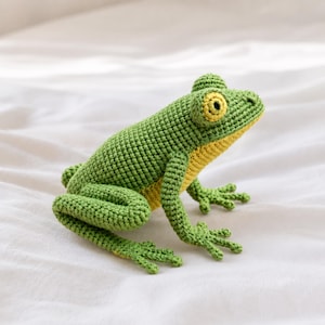 May include: A handcrafted crochet frog toy featuring a textured green body with a contrasting yellow underside and belly. The frog has prominent, round yellow eyes with dark pupils and detailed webbed feet, all created with tight yarn stitches.