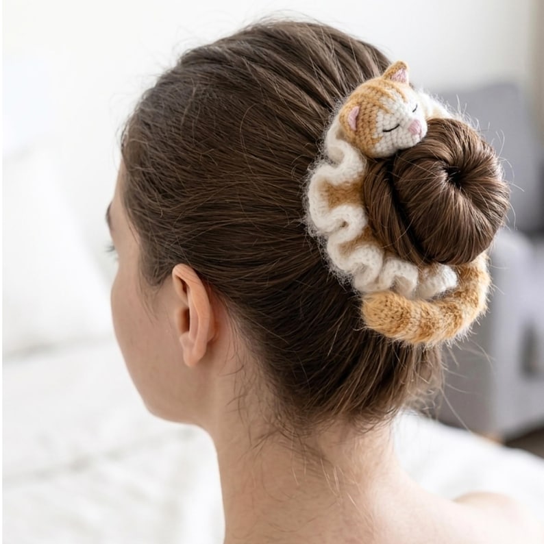 May include: A crocheted hair scrunchie featuring a sleeping cat design. The scrunchie is white and tan, with a matching cat figure. The cat is tan and white, with closed eyes. The scrunchie is wrapped around a bun.