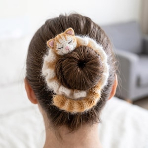 Könnte beinhalten: Handgefertigtes Häkel-Haaraccessoire mit einem schlafenden Katzendesign. Die Katze ist orange und weiß, mit geschlossenen Augen und rosa Akzenten. Das Haaraccessoire ist um einen Dutt gewickelt, mit einem cremefarbenen und orangefarbenen Streifenmuster.